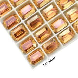May include: A close-up of a tray of 14x10mm emerald-cut rhinestones. The rhinestones are a warm amber color with a slight iridescent shimmer. The tray is white and has a grid pattern.