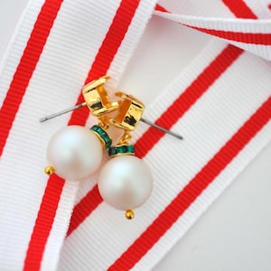 May include: A pair of pearl earrings with gold-colored accents and green gemstones. The earrings are set against a white ribbon with red stripes, creating a striking contrast. The earrings have a classic, elegant design.