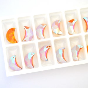 May include: A white plastic tray with 12 crescent moon shaped iridescent glass rhinestones. The rhinestones have a rainbow effect and are arranged in rows. One rhinestone is orange.
