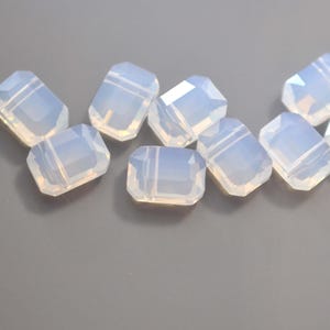 May include: Close-up of several opalescent, rectangular beads with faceted edges. The beads have a translucent, milky white color and are arranged on a gray surface. These beads are suitable for jewelry making.