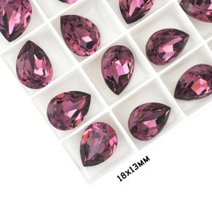 May include: Close-up of a tray of faceted, teardrop-shaped crystals in shades of purple. The crystals are 18x13mm in size and are arranged in a grid pattern. The crystals have a high-gloss finish and reflect light. The text "18x13MM" is visible.