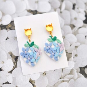 Peut inclure: Une paire de boucles d'oreilles pendantes avec un fond blanc et des motifs de fleurs bleues, vertes et violettes. Les boucles d'oreilles sont en pâte polymère et ont des crochets dorés.