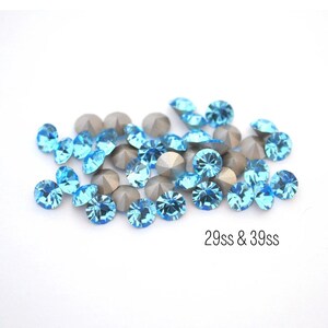 May include: A pile of light blue rhinestones with silver settings. The rhinestones are scattered on a white background. The text "29ss & 39ss" is visible below the rhinestones.