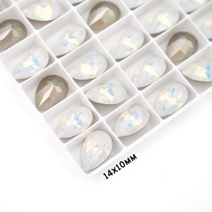 May include: A close-up of a white plastic container filled with iridescent white teardrop-shaped rhinestones. The container is labelled "14x10mm".