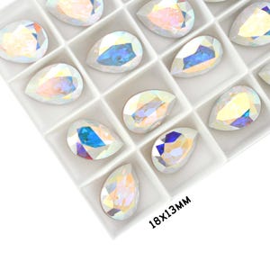 May include: A close-up view of a white plastic tray filled with iridescent teardrop-shaped rhinestones. The tray is labeled "18x13mm".
