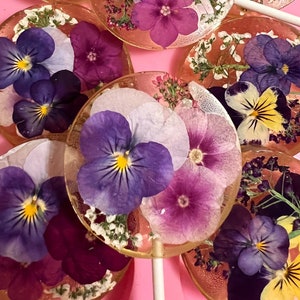 May include: Assortment of colorful lollipops with pressed flowers, including purple, yellow, and white pansies. The lollipops are on a pink background.
