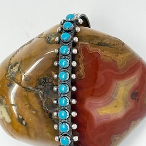 May include: A silver bracelet with turquoise stones. The bracelet features a row of bright blue turquoise cabochons set in individual silver settings. Small silver beads accent the sides of the bracelet. The bracelet is displayed on a brown and red stone.
