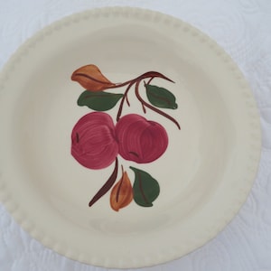 May include: Cream-colored ceramic plate with a scalloped edge. The plate features a hand-painted design of two red apples with green leaves and brown stems. A charming piece for kitchen decor.