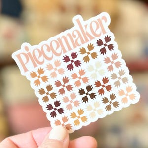 May include: A square sticker with a scalloped edge featuring the word "Piecemaker" in a light peach font at the top. Below the text is a repeating grid pattern of small, four-pointed star shapes in various shades of brown, tan, cream, and peach.