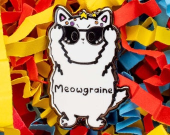 May include: White enamel pin of a cat wearing sunglasses with the text "Meowgraine" on the front. The pin is surrounded by colorful paper shreds.