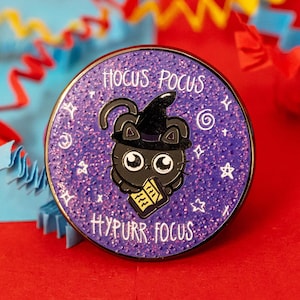 Op de afbeelding: Een zwarte kat met een heksenhoed en een boek in zijn pootjes. De kat is omringd door paarse glitter en sterren. De tekst "Hocus Pocus Hyppurr Focus" staat rondom de kat.