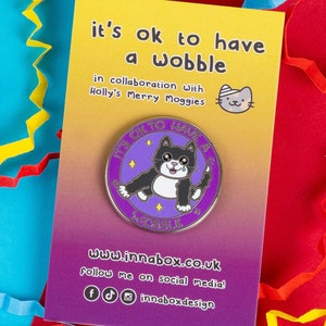 以下が含まれることがあります： 紫と金のエナメルピン。黒と白の猫が「It's ok to have a wobble」という文字の入った円の中に描かれています。ピンは、「it's ok to have a wobble in collaboration with Holly's Merry Moggies」という文字とウェブサイトのアドレスが書かれたカードの上に置かれています。