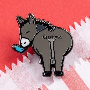 May include: Black enamel pin of a donkey with the word "Assthma" written on its back. The donkey is wearing a blue inhaler.