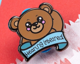 Puede incluir: Un pin de esmalte de dibujos animados de oso marrón con una pancarta azul que dice "Hearing Imbearred".
