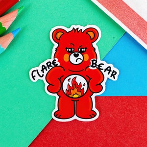 A sticker of a red bear looking grumpy with a flame on its tummy with the words FLARE BEAR on its shoulders it is on a green, red and blue background with pencils in in the corner of the shot.