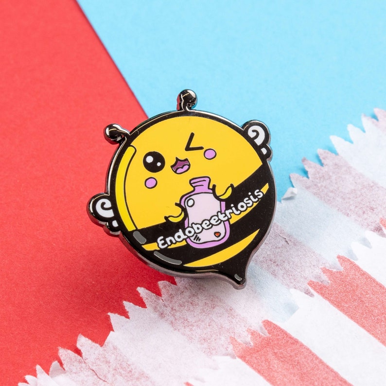 Endobeetriosis 2.0 Bee Enamel Pin - Endometriosis on red & blue card. A smiling bumble bee holding a pink hot waterbottle in its hands, across its middle reads endobeetriosis. The enamel pin is designed to raise awareness for endometriosis.