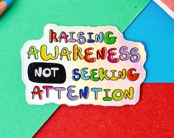 May include: A holographic sticker with the text "Raising Awareness Not Seeking Attention" in rainbow colors. The words are in a bubble font.