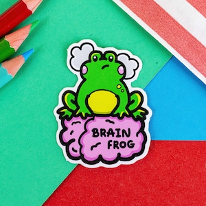 The Brain Frog Sticker - Brain Fog on a green, red & blue background with pencils. A confused frog with pink cheeks & 2 grey clouds above. Its sat on a pink brain with text that reads Brain Frog. Design was created to raise awareness for brain fog.