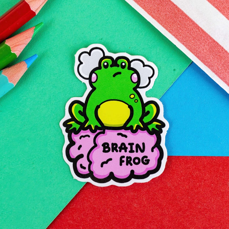 The Brain Frog Sticker - Brain Fog on a green, red & blue background with pencils. A confused frog with pink cheeks & 2 grey clouds above. Its sat on a pink brain with text that reads Brain Frog. Design was created to raise awareness for brain fog.