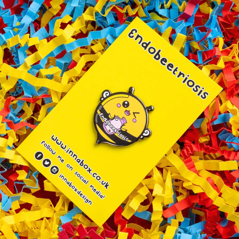 Endobeetriosis 2.0 Bee Enamel Pin - Endometriosis on yellow backing card laid on yellow, red & blue card. Smiling bee holding a pink hot waterbottle, across its middle reads endobeetriosis. Enamel pin is designed to raise awareness for endometriosis.