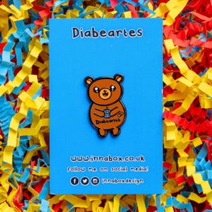 Diabeartes Enamel Pin - Diabetes on blue backing card on yellow, red & blue card. Smiley brown bear with a blood glucose reader & a plaster on its arm, across its tummy reads diabeartes in black. Raising awareness for type 1 & type 2 diabetes.