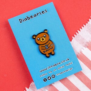 Diabeartes Enamel Pin - Diabetes on blue backing card on red & blue card. Smiley brown bear holding a blood glucose reader with a plaster on its arm, across its tummy reads diabeartes in black. Raising awareness for type 1 diabetes & type 2 diabetes.