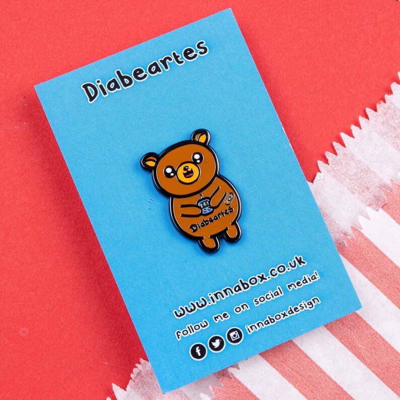 Diabeartes Enamel Pin - Diabetes on blue backing card on red & blue card. Smiley brown bear holding a blood glucose reader with a plaster on its arm, across its tummy reads diabeartes in black. Raising awareness for type 1 diabetes & type 2 diabetes.