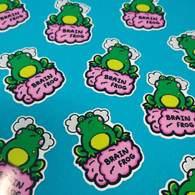 Brain Frog Sticker - Brain Fog, multiple frog stickers on a blue background. A confused frog with pink cheeks & 2 grey clouds above. Sat on a pink brain with text reading Brain Frog. Created to raise awareness for brain fog.