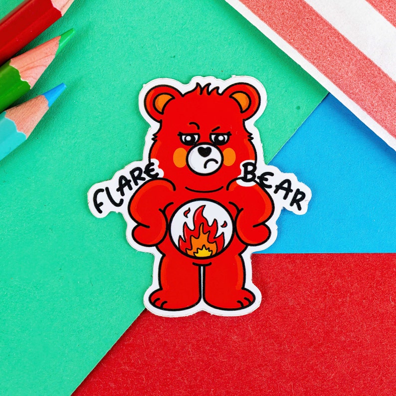 A sticker of a red bear looking grumpy with a flame on its tummy with the words FLARE BEAR on its shoulders it is on a green, red and blue background with pencils in in the corner of the shot.