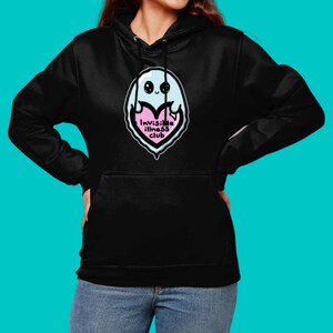 Invisible Illness Club Black Hoodie modelled on a blue background. It has a pastel blue smiling ghost holding up a pink heart with text reading invisible illness club. It has a front pocket & drawstring hood. Raising awareness for hidden disabilites.