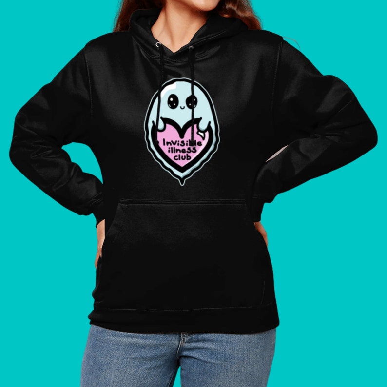 Invisible Illness Club Black Hoodie modelled on a blue background. It has a pastel blue smiling ghost holding up a pink heart with text reading invisible illness club. It has a front pocket & drawstring hood. Raising awareness for hidden disabilites.