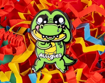 May include: Green alligator pin with a sad expression, wearing a white shirt with the word "Alligies" printed on it. The pin is surrounded by colorful paper shreds.