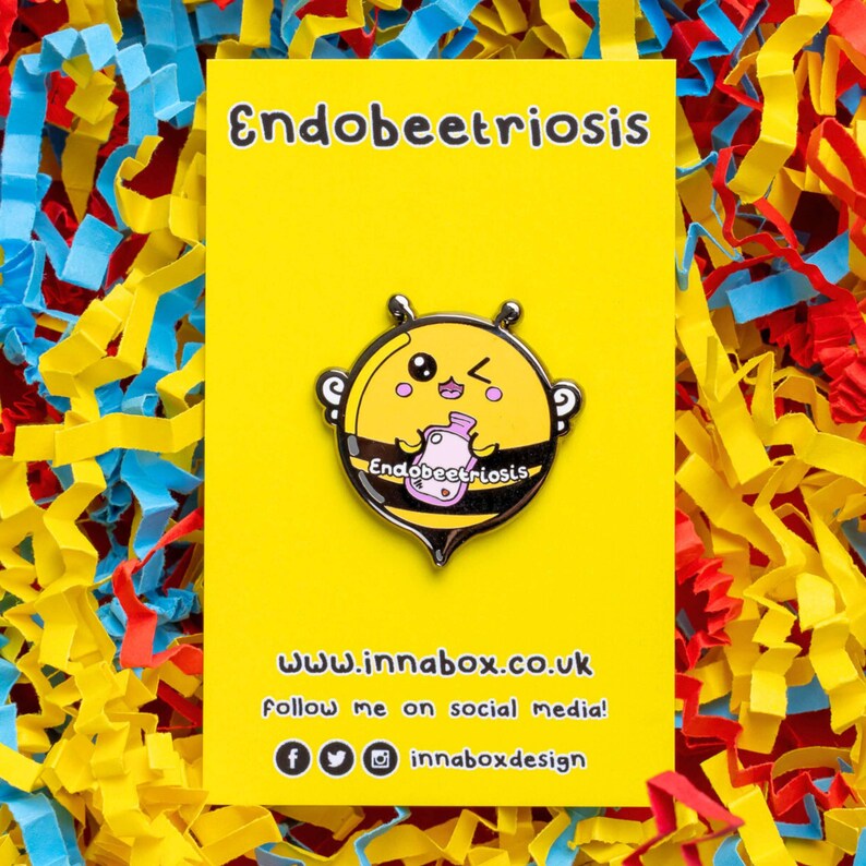 Endobeetriosis 2.0 Bee Enamel Pin - Endometriosis on yellow backing card laid on yellow, red & blue card. Smiling bee holding a pink hot waterbottle, across its middle reads endobeetriosis. Enamel pin is designed to raise awareness for endometriosis.