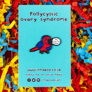 Puede incluir: Un pin de esmalte azul y rojo de un loro sosteniendo un bocadillo que dice "Polly wants new OVARIES". El pin está sobre un fondo azul con el texto "Pollycystic ovary syndrome" en la parte superior y "www.innabox.co.uk" en la parte inferior.