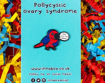 May include: A blue and red enamel pin of a parrot holding a speech bubble that says "Polly wants new OVARIES". The pin is on a blue background with the text "Pollycystic ovary syndrome" at the top and "www.innabox.co.uk" at the bottom.
