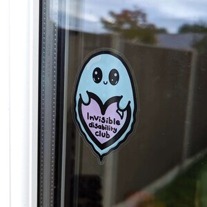 Invisible Disability Club Ghost Window Cling on a house window. Pastel blue smiling Ghost window sticker holding a pink heart with text reading invisible disability club. Raising awareness for hidden disabilities and invisible illnesses.