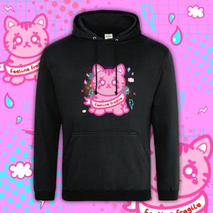 Black Feeline Fragile Cat Hoodie by Innabox featuring a kawaii pink cat illustration with emotional expression and pastel accents. Disability awareness clothing designed to support those with chronic illness and hidden disabilities. Soft, cosy hoodie with bold, cute graphic print.