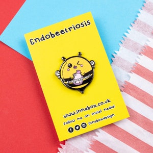 Endobeetriosis 2.0 Bee Enamel Pin - Endometriosis on yellow backing card laid on red & blue card. Smiling bumble bee holding a pink hot waterbottle, across its middle reads endobeetriosis. Enamel pin is designed to raise awareness for endometriosis.