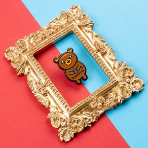 Diabeartes Enamel Pin - Diabetes on red & blue card in a gold frame. Smiling brown bear holding a blood glucose reader with a plaster on its arm, across its tummy reads diabeartes in black. Raising awareness for type 1 diabetes & type 2 diabetes.