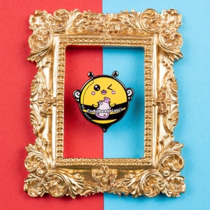 Endobeetriosis 2.0 Bee Enamel Pin - Endometriosis on red & blue card in a gold frame. A smiling bumble bee holding a pink hot waterbottle in its hands, across its middle reads endobeetriosis. Designed to raise awareness for endometriosis.