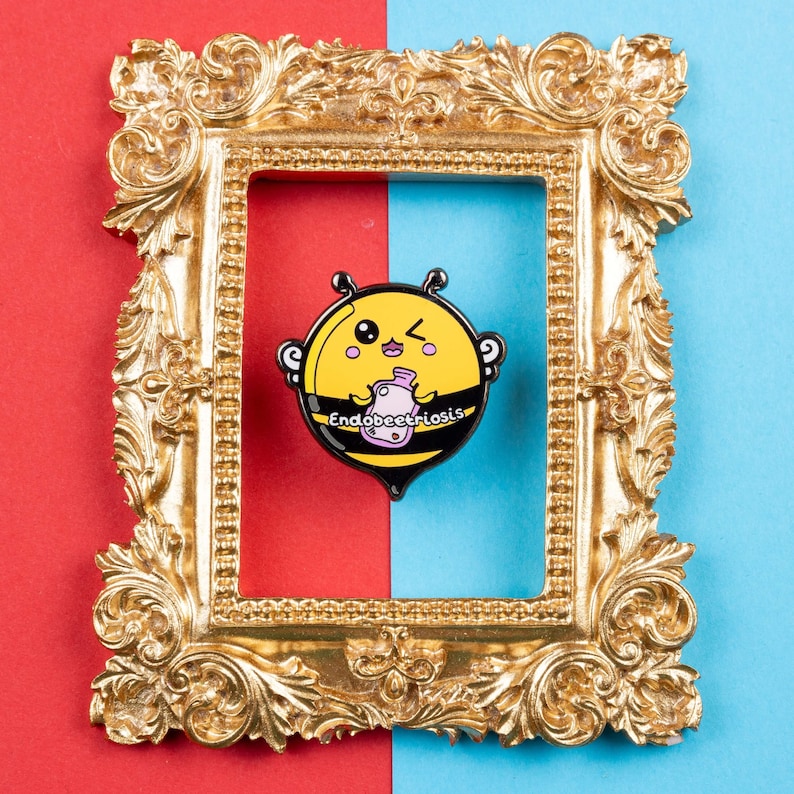 Endobeetriosis 2.0 Bee Enamel Pin - Endometriosis on red & blue card in a gold frame. A smiling bumble bee holding a pink hot waterbottle in its hands, across its middle reads endobeetriosis. Designed to raise awareness for endometriosis.