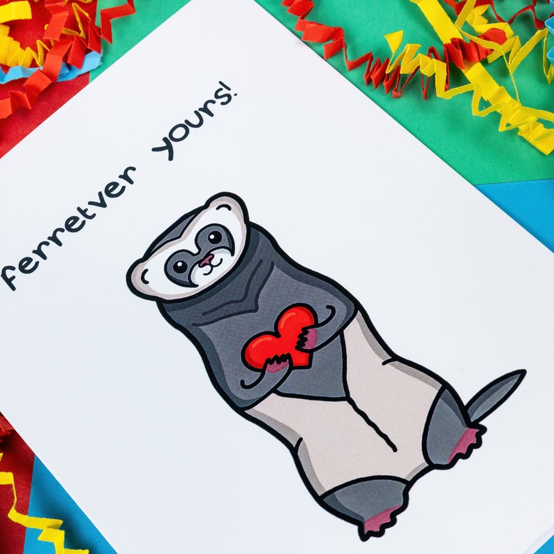 The Ferretver Yours Valentines Day Love Card on a red, blue and green background with red, yellow and blue crinkle card confetti. A white a6 greetings card with a drawing of a happy ferret holding a red heart saying ferreter yours on the front.