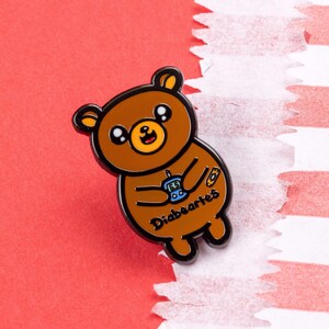 Diabeartes Enamel Pin - Diabetes on red card in a gold frame. A smiling brown bear holding a blood glucose reader with a plaster on its arm, across its tummy reads diabeartes in black. Raising awareness for type 1 diabetes & type 2 diabetes.