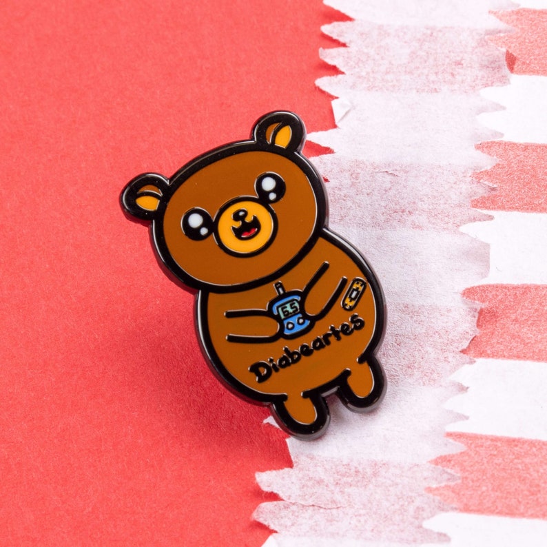 Diabeartes Enamel Pin - Diabetes on red card in a gold frame. A smiling brown bear holding a blood glucose reader with a plaster on its arm, across its tummy reads diabeartes in black. Raising awareness for type 1 diabetes & type 2 diabetes.