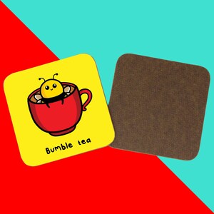 The Bumble Bee Tea Coaster on a red and blue background. The yellow wooden coaster features a cute bumble bee popping out a red cup of tea with black text underneath reading bumble tea, on the right is the wooden underside of the coaster.