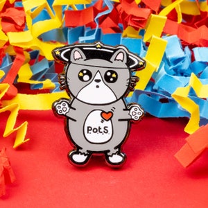 May include: A grey enamel pin of a cat wearing a star-shaped hat. The cat has large eyes and a heart on its chest that says "Pot.S."