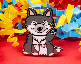 May include: A black and white enamel pin of a cartoon husky dog with a lightning bolt above its head. The dog is wearing a white collar with the text "TMJ" on it.