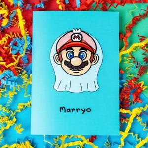 Marryo - Mario Engagement Wedding Card on red, blue & green card with crinkle card confetti. Blue a6 congratulations card of a smiling nintendo mario character head wearing a white bridal wedding veil, underneath him is black text reading Marryo.