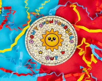 May include: A silver glitter sticker with a yellow sun with a smiling face and the text "Don't Burn Yourself Out" in rainbow colors.