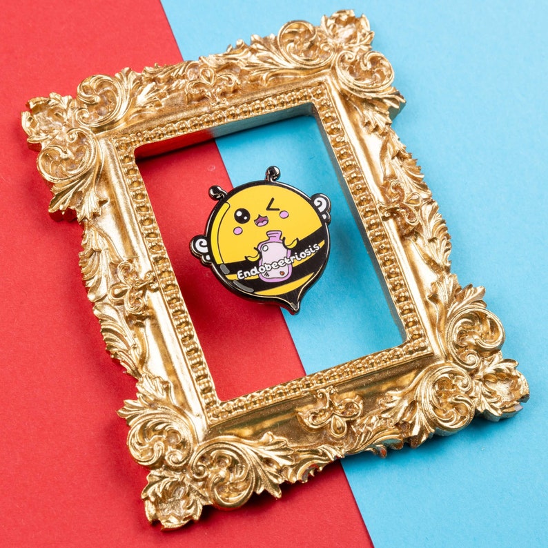 Endobeetriosis 2.0 Bee Enamel Pin - Endometriosis on red & blue card in a gold frame. A smiling bumble bee holding a pink hot waterbottle in its hands, across its middle reads endobeetriosis. Designed to raise awareness for endometriosis.